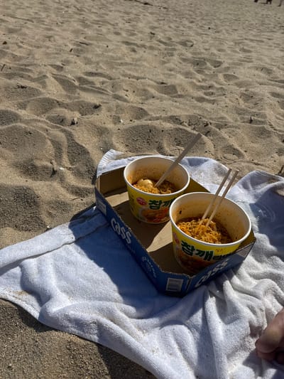Two cups of ramen, each with chopsticks, sit in a cardboard beer carrier on a white towel. The scene is on a sandy beach, presumably in Toseong-myeon, South Korea. A person's foot is visible on the towel. The ramen appears to be partially eaten. The towel is on the sand.