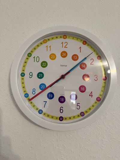 A close-up, eye-level, frontal shot shows a white, circular clock mounted on a textured, off-white wall. The clock face displays numbers from 1 to 60, with numbers 1 through 12 in a larger font and a secondary ring of numbers from 1 to 60 around the perimeter. There are colored circles highlighting certain numbers, such as 22, 20, 19, 18, 17, 16, 15, 14, 13, 24, 50, 45, 40, and 35. The clock has a red hour hand pointing to the number 7, a black minute hand pointing to the number 11, and a blue second hand pointing to the number 37. The clock face also features the brand name "hama". The wall appears to be an interior wall, possibly in a room within a residential or educational setting. There is a faint reflection on the clock face, suggesting the presence of light sources in the environment. The specific location in Velbert, Germany, is not identifiable from the provided image.