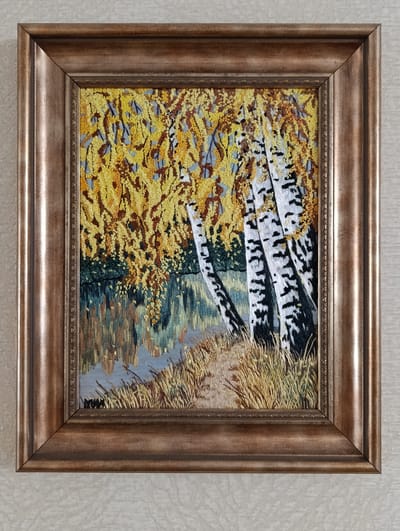 The image shows a framed embroidery of birch trees by a body of water. The trees have white trunks with black markings and are surrounded by yellow foliage. Grass grows along the edge of the water. The frame is a dark gold color with ornate detailing. The background is a light beige textured wall. There is no evidence to indicate its location in Barvinok, Ukraine.