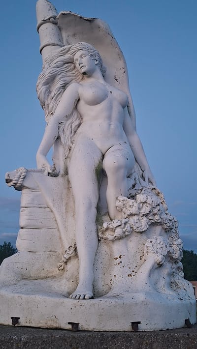 The image shows a full-body, white, weathered statue of a partially nude female figure situated on a stone base. The statue is positioned outdoors against a clear blue sky with hints of clouds near the horizon. The female figure is depicted with long, flowing hair, a bare torso, and is seated on what appears to be part of the sculpture's base, which incorporates abstract, wave-like forms and a rope detail. The statue exhibits signs of age and weathering, with some green algae and brown discoloration visible on its surface. The base of the statue has metal fixtures protruding from it. In the background, out-of-focus trees and structures are visible, suggesting an outdoor urban or park-like setting. This statue is located in Paldiski, Estonia.