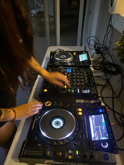 A DJ station features three Pioneer devices: two CDJ-2000 Nexus multiplayers on either side and a DJ mixer in the middle. Wires are visible above the equipment. A person's hands are manipulating the mixer, while long brown hair obscures the lower portion of the image. A tattoo is visible on one of the person's arms, adorned with a bracelet. The lit screens and jog wheels of the CDJs are a focal point, indicating active use.