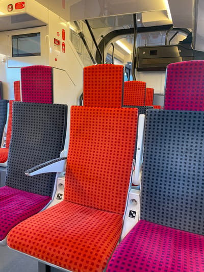 The image depicts the interior of a modern, empty passenger train carriage, specifically located at or departing from Plaisir Les Clayes station in Plaisir, France, with "Paris Montparnasse" indicated as the destination on an overhead digital display. The carriage features numerous rows of fabric-upholstered seats in a repeating pattern of small squares. Seat colors alternate between a bright red-orange, a dark grey, and a magenta/fuchsia. Some seats have white armrests. White panels between seat pairs contain a European-style power outlet and a USB charging port, with labels indicating 230V~, 50Hz, 750VA max, and 5V, 2A respectively. The interior walls and ceiling panels are predominantly white or light grey, with integrated linear lighting. A red "Alarme" (Alarm) button and other control panels are visible on the wall near the display screen. No individuals are present in the scene.