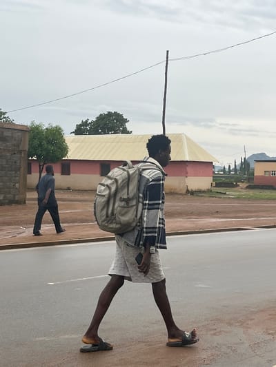 In Sabojibi, Nigeria, the image depicts an outdoor scene with two individuals. The primary subject is a young man walking on a paved surface, possibly a road or pathway, carrying a light-colored backpack. He is wearing a checkered jacket over a white shirt and patterned shorts. His footwear appears to be dark slip-on sandals. To his left, a building with a light-colored roof and red and beige walls is visible. Further in the background, more buildings and trees are scattered, with a hill in the distance.

Behind the main subject, another person is walking away from the camera. This individual is wearing a dark blue shirt and dark pants. They are seen near a brick structure and some greenery.

The sky is overcast, suggesting it might be a cloudy day, possibly with rain or having recently rained, as indicated by some wet patches on the ground. A thin wire stretches across the upper portion of the image, supported by a wooden pole. The overall atmosphere is casual and everyday, capturing a moment of transit or daily life in the depicted location. There is no visible text in the image.