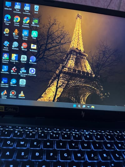 A close-up shot reveals a laptop with a backlit keyboard. The screen displays a wallpaper featuring the Eiffel Tower in Paris, shrouded by silhouetted trees against a golden sky. Desktop icons for various applications are visible, including Google Chrome, Microsoft Edge, Adobe After Effects, and several gaming applications. The bottom right of the screen indicates "Full HD 144Hz." The arrangement suggests a blend of professional and leisure use.
