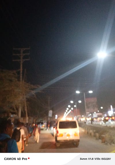A blurry night scene captures a street lined with people and vehicles. A white van is prominently positioned in the foreground, its taillights visible. Streetlights illuminate the road, casting a glow over the scene. On the left, pedestrians walk along the sidewalk. A utility pole stands tall amidst bare trees. The overall image has a grainy texture due to low lighting conditions. The camera model and settings are visible at the bottom of the frame.
