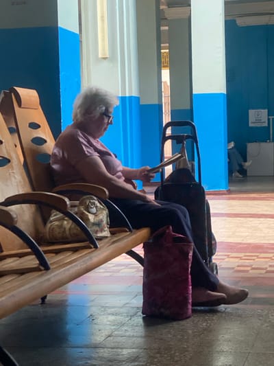 In Romodan, Ukraine, an elderly woman with white hair and glasses sits on a wooden bench in what appears to be a waiting area. She is wearing a pink shirt and dark pants, and her feet are bare. Beside her, to her right, is a dark rolling suitcase with a handle. A small, ornate bag rests on the bench to her left. A red bag is on the floor in front of the bench. The background features bright blue walls and large white columns, with a tiled floor. In the distance, another person is visible, sitting in a chair. The scene conveys a sense of waiting or transit.