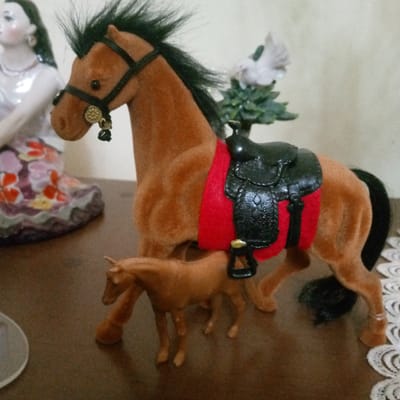 This image displays a still life arrangement of figurines, likely on a table, with a blurred background suggesting an indoor setting. The primary subjects are two toy horses, one larger and one smaller, with a saddle and red saddle blanket on the larger horse. The horses appear to be made of a fuzzy material, with black manes and tails. The larger horse has a black bridle with decorative gold accents.

To the left of the horses is a porcelain figurine of a woman wearing a shimmering, ornate dress patterned with colorful floral designs. Her facial features are delicate, and she is adorned with earrings and a necklace.

In the background, behind the larger horse, is a small decorative tree with a white figurine of a bird perched on its branches. The overall scene is static, with no activity taking place. The lighting appears to be artificial, casting subtle shadows and highlighting the textures of the figurines. The presence of lace trim on the right side of the image suggests it may be part of a tablecloth or decorative runner. There is no visible text in the image. The context provided indicates a location in Ashtarak, Armenia, but this information is not visually present in the image itself.