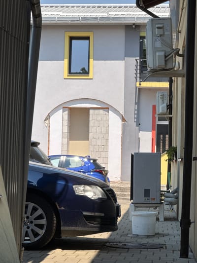 This image depicts an outdoor scene, likely a residential or commercial building's exterior, in Suceava, Romania. The orientation of the image is vertical.
In the foreground, a dark-colored vehicle, possibly a van or SUV, is partially visible. Its front wheel, with a silver alloy rim and a Volkswagen logo, is clearly seen. To the left of this vehicle, another blue car is parked.
The background reveals the facade of a building with a light gray and white color scheme. There is a rectangular window with a bright yellow frame, reflecting the blue sky and trees. Above the window, an outdoor air conditioning unit is mounted on the wall, with pipes and electrical wiring connected to it. Further to the right, a large gray electrical cabinet or utility box is visible, and next to it, a white bucket. The building's architecture includes arched openings and walls constructed with light-colored bricks. The ground appears to be paved with rectangular stones or bricks.
The lighting suggests it is daytime, with clear sunlight casting shadows, indicating fair weather. The overall impression is of an organized, perhaps somewhat utilitarian, outdoor space adjacent to a building.
