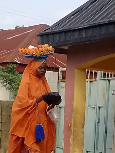 A young Black woman wearing an orange hijab and robe is walking outdoors in Gbaza, Nigeria. She is carrying a tray of eggs balanced on her head, and has a black object in her hands, possibly a bird. There is a blue pouch hanging from a chain on her robe. In the background, there are buildings with corrugated metal roofs and a light green metal gate. The weather appears to be overcast.