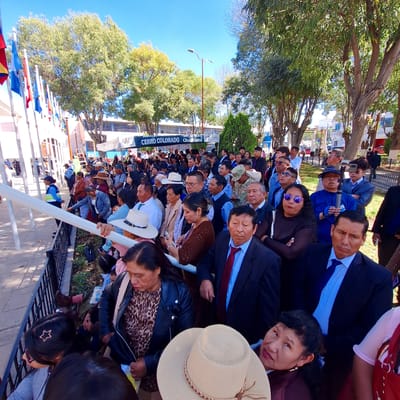 A large group of people, dressed in a mix of casual and formal attire, are gathered outdoors in what appears to be a public space. The banner of Cerro Colorado is visible. Most of the people are seated and facing in the same direction, suggesting they are watching an event or presentation. Trees provide shade over the scene. Several individuals are wearing hats, and some are wearing sunglasses. The location is Cerro Colorado, Peru.
