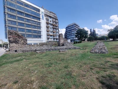 The image shows an outdoor scene in Thessaloniki, Greece. In the foreground, there is a grassy area with some remnants of ancient stone structures, appearing to be ruins. Two long, flat, rectangular stones are visible in the grass. Behind the ruins, two modern buildings rise up. The building on the left is a tall, white structure with a facade composed of many glass windows and vertical metal elements. A green logo with blue lettering, possibly "EMG", is visible on its side. To the right of this building is a lighter-colored residential building with multiple balconies. Further in the background, another contemporary building with a curved facade and blue accents can be seen. Trees are scattered throughout the scene, and a clear blue sky with scattered white clouds is overhead. The overall impression is a juxtaposition of ancient history with modern urban development.