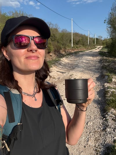 A selfie shows a person wearing sunglasses, a black baseball cap, a silver necklace, a black shirt, and a blue backpack, holding a black cylindrical object in their right hand. The person has a silver nose ring. The background reveals a dirt path surrounded by vegetation. White poles, likely for electrical or communication lines, are visible in the distance along the path. The scene appears to be outdoors in Podbadac, Montenegro.