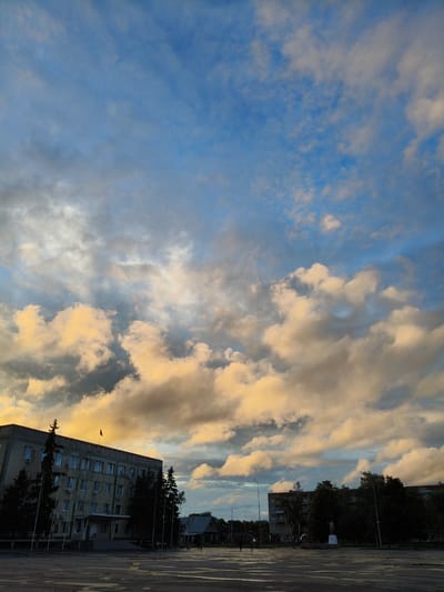 A wide-angle, eye-level shot captures an overcast sky filled with dramatic clouds tinged with yellow and orange hues, suggesting either sunrise or sunset. Beneath the expansive sky, a city square is visible. On the left, a large, multi-story building with numerous windows and air conditioning units dominates the scene. A flag flies from its rooftop. Several dark, slender trees are planted in front of this building.
Further into the square, to the right, a statue of a person is visible, surrounded by trees. The ground in the foreground appears wet, reflecting the sky and surrounding structures, indicating recent rain or damp conditions. A few scattered figures are present in the distance, walking across the square. The overall impression is one of a quiet, possibly post-rain, urban landscape under a dynamic sky. The specific location is the city of Petrovsk, Russia.