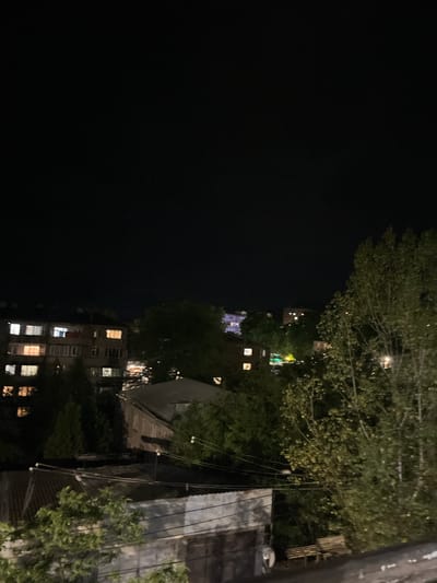 This media file depicts a nighttime scene in Gladzor, Armenia. The view is from an elevated perspective, looking down and across a cityscape that is partially obscured by trees and foliage. Several residential buildings are visible, characterized by illuminated windows indicating occupancy and activity within. The architecture suggests an urban or semi-urban environment. The time of day is clearly night, with a dark sky and artificial lighting from the buildings and possibly streetlights providing illumination. The presence of trees suggests a temperate climate with foliage still on the branches, though the exact season is not definitively indicated. There are no people or specific objects that are the primary focus, rather the overall scene of a residential area at night. The overall mood is one of quiet observation, capturing the nocturnal ambiance of the location. No text is visible in the media file.
