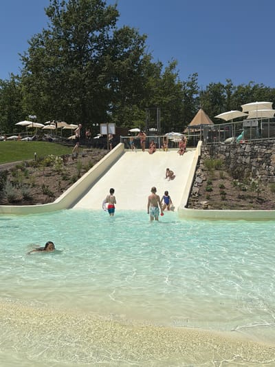 In Cavriglia, Italy, this image depicts a clear summer day at a water park. A wide, white water slide dominates the scene, curving down into a shallow pool filled with crystal-clear turquoise water. Several children are actively engaged in water activities. On the slide, a young child is midway down, and a few children are at the top or sliding down in pairs. In the pool, a young boy in red and blue swim trunks is standing with a circular float, while another boy in blue shorts stands further back. To the left, a girl with long dark hair is swimming in the shallow water. Behind the slide, a group of people are gathered, some sitting on the edge of the slide, while others are standing and observing. Trees and lush green grass surround the pool area, with lounge chairs and umbrellas visible in the background, suggesting a recreational area. The sky is a vibrant, cloudless blue.