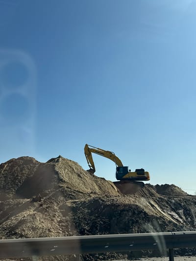 A yellow and black Sumitomo excavator is positioned on a large, light brown earthen mound under a clear, cloudless blue sky in Aiuta, Russia. The excavator's boom and bucket extend downwards into the primary dirt pile. Further, uneven mounds of earth, exhibiting varied tones, are visible around the excavator. A dark gray metal guardrail runs horizontally across the bottom of the frame, slightly out of focus. Two faint, blurred ring-shaped reflections are present in the upper-left quadrant of the image. No individuals or specific actions are discernible.