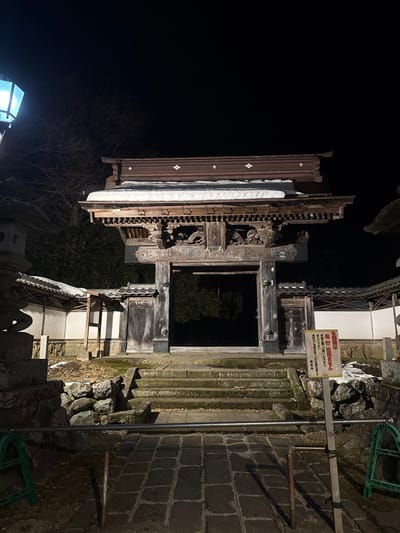 The image depicts a snow-covered traditional Japanese temple gate at night in Yamanouchi, Japan. A multi-tiered wooden gate, centrally positioned, features intricate carvings on its lower structure, and its dark roof is topped with a layer of snow. A paved stone pathway in the foreground leads to a series of moss-covered stone steps ascending to the gate's entrance. A horizontal metal pipe runs across the lower pathway. To the left, a stone lantern and a white plaster wall with a tiled roof are visible, alongside bare tree branches against a dark sky. To the right, a similar white plaster wall with a tiled roof is present, accompanied by a white rectangular sign displaying Japanese characters in red and black. Patches of snow are scattered on the ground, steps, and surrounding structures, illuminated by artificial light which casts subtle shadows. No individuals are present in the scene.