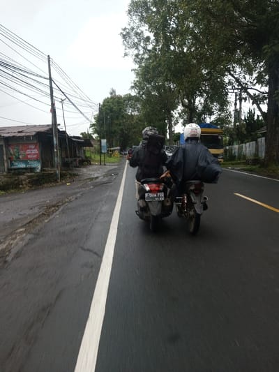 The image depicts an outdoor scene from a slightly elevated perspective, looking forward on a wet, two-lane asphalt road under an overcast sky.
Two individuals are riding motorcycles in the foreground, moving away from the viewer. The person on the left is on a dark-colored scooter, specifically a model resembling a Yamaha NMAX, with a rear license plate displaying "F 5050 FEU" and "01-23" below. This rider wears a grey full-face helmet, a dark jacket, and carries a black backpack. The person on the right is on a lighter-colored motorcycle, likely an underbone type, and wears a white full-face helmet and a dark, billowy rain poncho or jacket.
To the left of the road, a dirt shoulder parallels the asphalt. Beyond it, a row of dilapidated, single-story structures with corrugated metal roofs are visible, appearing to be small shops or stalls. A prominent red and white sign on one structure partially displays Indonesian text including "JUAL PULSA" (Sell Credit) and "INDOSAT." A wooden utility pole stands in front of these structures, densely covered with numerous black electrical or communication wires. The ground near the structures is unpaved and wet.
To the right of the road, dense green trees line the roadside, continuing into the mid-ground. A light-colored fence or wall is partially visible beneath the trees. In the distance, a yellow and blue truck is visible on the road, also moving away from the camera's perspective. The sky is a uniform bright grey, indicative of heavy cloud cover.
Regarding the requested location, the scene is described as being in Serpang Shalli, Bhutan. However, visual evidence from the image, specifically the "F 5050 FEU" license plate and the Indonesian text "JUAL PULSA" on a roadside sign, strongly indicates the location is in Indonesia, not Bhutan.
