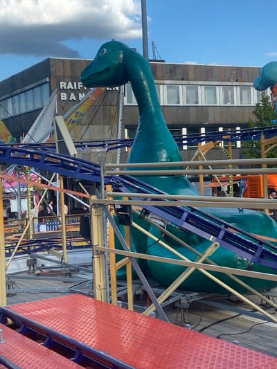The image shows a large, green dinosaur amusement park ride in the foreground, with a roller coaster track winding around it. In the background, a building with the words "RAIFFEISEN BANK" is visible. The sky is blue with some clouds. The scene appears to be a carnival or amusement park in Baunatal, Germany.
