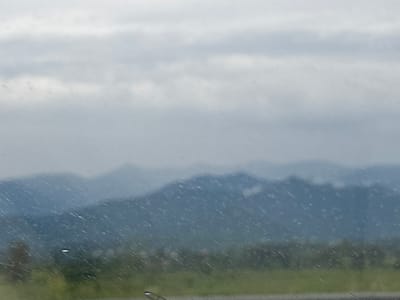 The image depicts a landscape scene, likely viewed through a wet window, given the streaks and droplets visible. In the foreground, a strip of green land is present, with faint hints of foliage. Beyond this, a series of undulating, blue-toned mountain ranges recede into the distance. The sky is overcast with layers of gray clouds, suggesting a cloudy or possibly rainy day. The overall impression is one of a hazy, muted natural environment. The scene is devoid of any visible people or specific structures that would definitively place it within the city of Kobuleti, Georgia, although the atmospheric conditions and mountainous backdrop are consistent with regions surrounding coastal towns.
