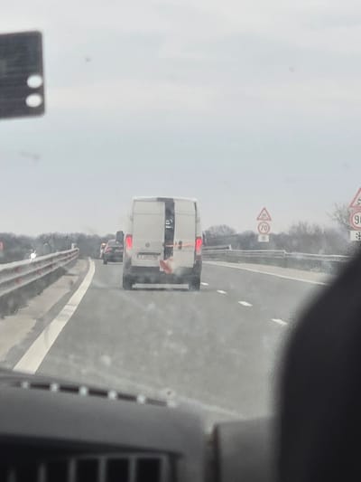 The image depicts a white panel van traveling away from the viewer on a two-lane asphalt highway in the vicinity of Opalchenets, Bulgaria, under an overcast sky. The van, possibly a Mercedes-Benz Sprinter or similar model, features a distinct vertical black stripe on its rear right door. A Bulgarian license plate, partially visible as "PA 9557 MH", is affixed to its rear. A red object, appearing to be fabric or plastic, protrudes from the lower right rear of the van, near the taillight. Further ahead in the same lane, a dark-colored sedan is visible.
The highway features clearly marked lanes with dashed white lines separating them and a solid white line defining the left shoulder. A metal guardrail runs along the left side of the road. On the right side of the highway, multiple road signs are visible: a triangular warning sign with a vehicle skidding, a circular speed limit sign indicating "70", and another circular sign displaying "90" with a snowflake symbol below it, likely indicating a winter speed limit or advisory. The photograph is taken from inside a vehicle, looking through a windshield, with the top of a dashboard and dark interior elements visible at the bottom and right edges of the frame. The overall light is even, consistent with daytime overcast conditions.