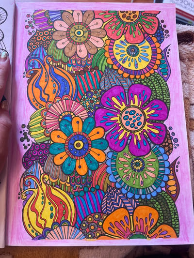 The image shows a close-up of a coloring book page filled with intricate, colorful floral designs. The artwork is a mandala-like composition featuring various stylized flowers, leaves, and abstract wavy patterns. The colors are vibrant, including shades of purple, pink, orange, yellow, green, blue, and brown, applied in a detailed, patterned manner. Some of the flowers have petal-like shapes with inner details, while others are depicted as circular motifs with radiating segments. The background of the coloring page is a soft pink, with some faint radiating lines suggesting a gentle glow. The overall impression is one of a decorative and artistic piece, likely created by hand with coloring markers or pencils. The image is likely taken from above, showing the coloring book lying flat. There is no visible text on the coloring book page itself. The background outside the coloring book is blurred, showing a textured brown surface, possibly fabric like a blanket or rug, and the edge of what looks like a book or other flat objects.
