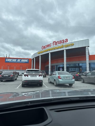 The image shows a view of the "Oasis Plaza" shopping and entertainment center in Beloyarsky, Russia, captured from inside a vehicle. Several parked cars are visible in the parking lot in front of the building. The license plates on the vehicles include E888PT 186, P320MO 186, and E905 PK 86. A sign for "Cinema de Luxe," a multi-screen 3D cinema, is visible on the building's facade. The sky is overcast.