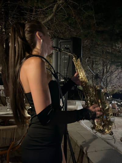 A woman with a high ponytail and long dark hair is playing a saxophone outdoors at night. She is wearing a black dress and black arm coverings. She is standing next to a black speaker. In the background, there are trees and string lights. There are tables and chairs visible, suggesting the location is a restaurant or outdoor dining area. This scene is set in the city of Pečurice, Montenegro.