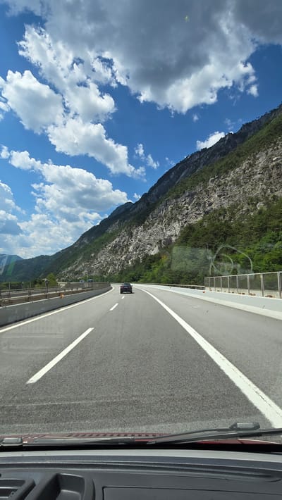 This is a point-of-view shot from inside a vehicle, likely a car, driving on a highway. The road is asphalt with clear white lane markings. A single dark-colored car is visible ahead, driving in the same direction. The highway curves to the left, bordered by a concrete barrier and a metal fence on the right. To the left of the road, there's another barrier and what appears to be construction or an embankment. A steep, rocky mountain slope covered in dense green trees dominates the right side of the image, extending upwards towards a bright blue sky filled with fluffy white and gray clouds. The scene is set in a mountainous region, consistent with the geography of Pontebba, Italy, known for its proximity to the Alps. The lighting suggests it is daytime, likely late morning or early afternoon, with sunlight casting shadows.