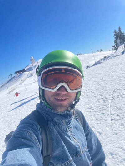 A male individual is depicted in a close-up, selfie-style photograph on a snow-covered mountain ski slope in Jahorina, Bosnia and Herzegovina. The subject wears a bright green ski helmet, white-framed ski goggles with orange reflective lenses, and a blue denim anorak jacket with a zippered collar. A black backpack strap is visible over his right shoulder. He has a beard and a slight smile, looking directly into the camera. The sky above is clear and blue. In the background, to the left, a ski lift structure ascends the snow-covered mountain. Further up the mountain, a prominent white dome structure, characteristic of an observatory or radar station, is visible at the peak. A small figure in a red jacket is discernible skiing or snowboarding further down the slope to the left. Snow-covered evergreens line a ridge on the right side of the background. Ski tracks are visible on the sunlit snow.