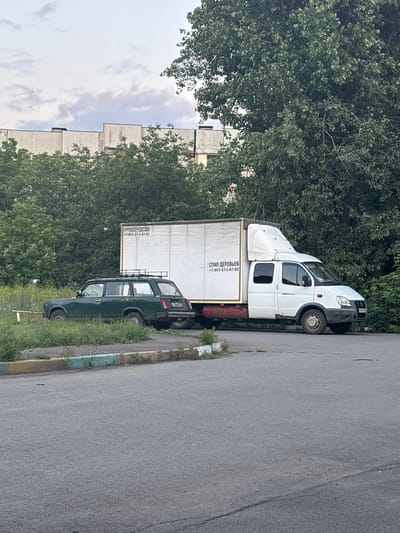 The image shows two vehicles parked on a paved surface next to overgrown grass and a curb. On the left is a dark green station wagon with a roof rack. To its right is a white box truck. The truck has text on its side, appearing to be in Cyrillic, with a phone number. In the background, there is a large tree with dense foliage and a multi-story building. The sky is cloudy. The scene is consistent with a residential or industrial area in Gukovo, Russia, given the presence of Cyrillic text on the truck and the style of the building. There are no people visible in the image.