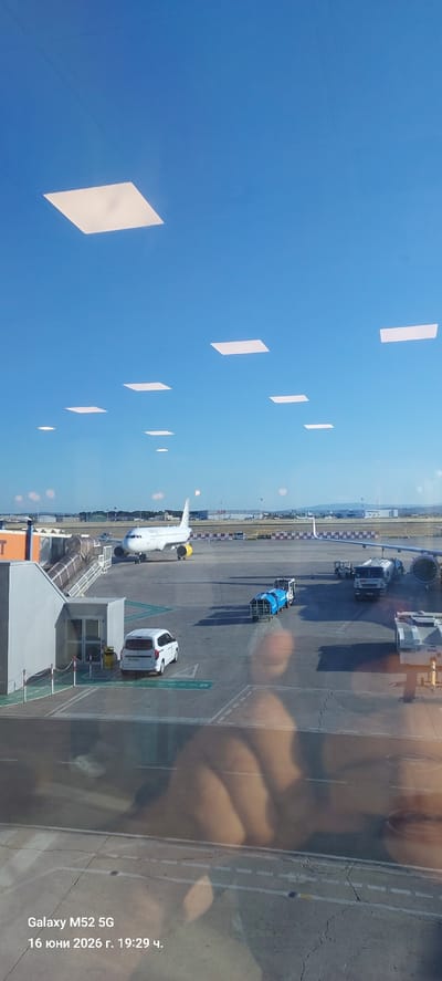 The image, taken at Valencia Airport (VLC) in Manises, Spain, depicts an airport tarmac scene under a clear blue sky. Several rectangular, white light panels are visible in the sky, likely part of the airport's interior ceiling reflected in the window. In the foreground, a white van is parked on the tarmac. A passenger jet, bearing the Vueling livery with yellow engine nacelles, is positioned on the tarmac, seemingly ready for boarding or disembarking, with a jet bridge connected to its fuselage. Several service vehicles, including a blue baggage tug and a white tanker truck, are present on the tarmac near the aircraft. In the background, airport buildings and infrastructure are visible, including hangars with "AIR NOSTRUM" signage. The lower portion of the image shows a reflection of what appears to be a person's hands and arms, as well as text indicating "Galaxy M52 5G" and a date/time stamp in Cyrillic script: "16 юни 2026 г. 19:29 ч." (June 16, 2026, 19:29).