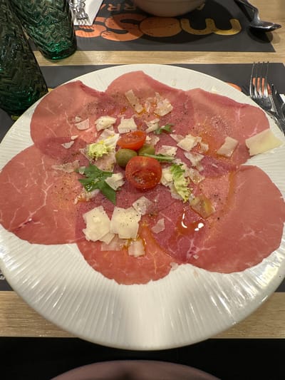 The image presents a top-down view of a carpaccio dish served on a white, textured plate, set in what appears to be a restaurant in Andorra la Vella, Andorra.
The main subject is a plate filled with thinly sliced red meat, likely beef carpaccio, arranged in an overlapping circular pattern. The meat is garnished with numerous toppings. Shaved or irregularly cut pieces of Parmesan cheese are scattered across the entire dish. Centrally placed are several halved red cherry tomatoes and a few small, green olives. Small sprigs of fresh rocket or lettuce leaves are also interspersed among the other ingredients. The dish is visibly drizzled with oil, which creates glossy highlights on the surface, and seasoned with finely cracked black pepper.
The white plate features a distinct ribbed or fluted texture along its rim, adding a decorative element. Surrounding the plate, parts of a dining setup are visible. To the left of the plate, a black placemat is present, featuring a large, stylized orange number "3" along with partial letters "OO" and "RR," possibly part of a restaurant's name or branding. A small piece of text "illa" can be seen near the bottom edge of the placemat. A wooden table edge runs alongside the placemat and the plate. In the bottom left, two dark green, highly textured glasses are partially visible. Cutlery, specifically a fork and a spoon, is visible near the top left edge of the plate, resting on the placemat. No people are visible, but the setup suggests an indoor dining experience ready for patrons.