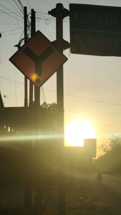 The image shows a fork in the road with a directional sign against a backdrop of a hazy sunset. A yellow diamond-shaped sign with a black Y-shaped symbol indicates a diverging path. Above it, a dark rectangular sign with white lettering is partially visible, reading "MONTASIK" and "BANDA AC". Telephone poles and wires are silhouetted against the sky. The sun, a bright orb, is low on the horizon, casting a strong glare and creating lens flare effects across the lower half of the image. Trees and some indistinct structures are visible in the background, obscured by the sunset's haze and glare. The scene appears to be on a road in or near Reuhattuha, Indonesia, given the style of the road signs which are characteristic of Indonesian signage. There are no discernible people in the frame.