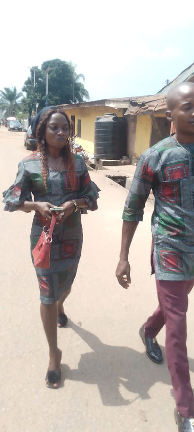 The image depicts an outdoor street scene in Ohafia, Nigeria, under bright daylight with strong shadows. Two individuals are walking on a light-colored paved road.
In the left foreground, a female subject walks towards the right. She has brown, wavy hair, partially covered by a black headscarf, and wears earrings and a necklace. She is dressed in a knee-length, V-neck dress with ruffled elbow-length sleeves, featuring a gray base with a repeating pattern of red and green geometric squares. She holds a white smartphone in her hands and carries a small red handbag on her left arm. A gold-colored bracelet is visible on her right wrist. She wears dark, open-toed sandals with a fluffy top. Her expression shows slightly pursed lips.
To her right, a male subject is partially visible, primarily from the chest down, also walking towards the right. He is bald. He wears a short-sleeved tunic with the same gray, red, and green geometric square pattern as the female's dress. He pairs this with maroon trousers and black dress shoes, which feature a gold-colored accent on the top.
The background shows an urban or semi-urban environment. A yellow-painted building with a corrugated metal roof is prominent, partially obscured by a large black cylindrical water tank situated on a raised concrete platform. Adjacent to it is another building with a rusty corrugated metal roof. Further down the road, two dark-colored cars are parked, and a motorcycle is visible. Two utility poles, potentially supporting streetlights, are present. Lush green trees, including palm trees, are visible against a bright, clear sky.