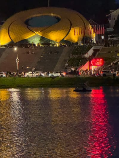 The image depicts a nighttime scene at Xuan Huong Lake in Da Lat, Vietnam. In the background, a large, illuminated golden structure resembling a lotus flower dominates the skyline. In front of this structure, a crowd of people is gathered. A row of red flags on poles is visible. To the right, a neon sign spelling "GO!" in bright red letters is displayed above a building entrance where more people are congregating. Cars are parked along the road in front of the gathering. The foreground of the image is filled with the dark water of Xuan Huong Lake, reflecting the lights from the background. A small boat is visible on the water. The overall atmosphere suggests a lively event or gathering at the lake.