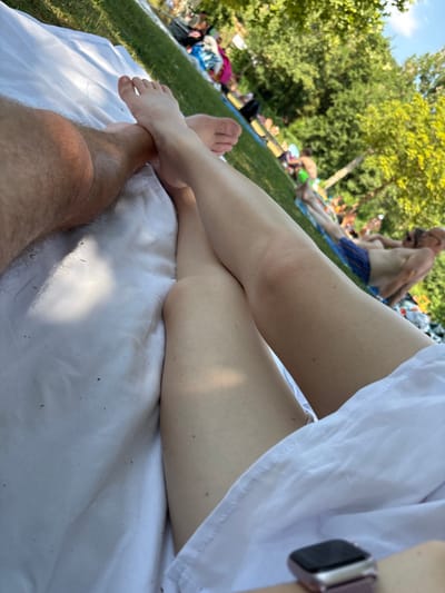 The image shows a close-up view of two people's legs and feet resting on a white blanket spread on a grassy area. In the background, other people are visible lounging on blankets and enjoying a sunny day outdoors. The setting appears to be a park or recreational area. The presence of trees and bright sunlight suggests an outdoor, possibly summer, environment. While the specific location in Bürgstadt, Germany is not explicitly identifiable from the visual elements, the scene depicts a relaxed gathering in a public outdoor space.
