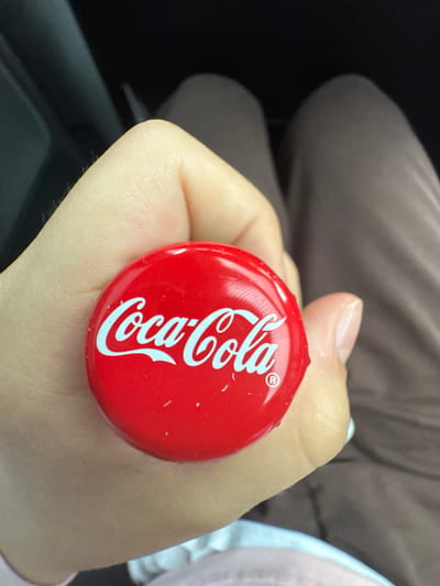 A person is holding a red Coca-Cola bottle cap in their hand. The cap has the white Coca-Cola logo on it, and a small registered trademark symbol. The background shows a dark interior, possibly a car, with the person's legs visible, wearing brown pants. The image is focused on the bottle cap and the hand holding it. The location is not determinable from the image itself.