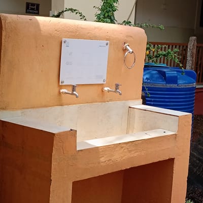 The image shows a public handwashing station. The station is constructed of painted stucco and features two faucets over a tiled basin. A sign with handwashing instructions is mounted above the faucets. To the right of the station is a blue water tank. The handwashing station is outdoors. The location is Marna, India. A sign reading "STRECHER" is located in the upper left corner of the image. A silver towel rack is mounted to the right of the handwashing instructions.