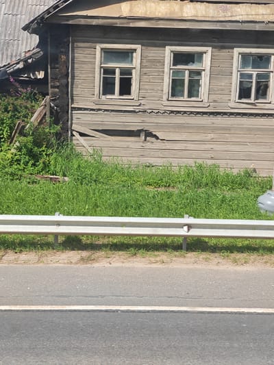 A weathered, wooden house stands partially obscured by overgrown grass and vegetation in Novaya Sitenka, Russia. The house has a dilapidated appearance with multiple broken window panes and exposed wooden beams. A metal guardrail runs horizontally across the foreground, separating the house and overgrown area from a paved road. The sky is not visible in the frame.