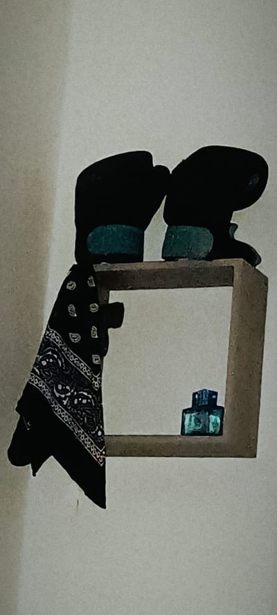 This is a close-up, eye-level, medium shot, of a shelf mounted on a light-colored wall. The shelf is made of a light brown wood and has a square opening. On the top of the shelf, on the left side, is a black bandana with white paisley patterns, hanging over the edge of the shelf. To the right of the bandana, on the top shelf, are two black, rounded objects with a teal-colored band around their base. These objects appear to be abstract sculptures. On the right side of the shelf, inside the opening, are two small, stacked, teal-colored objects. The overall lighting is dim and the image has a grainy texture. The location in the city of Winnenden, Germany, is not discernible from the image.