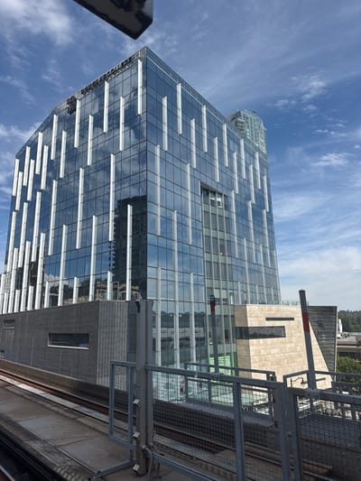 The image shows a modern glass-facade building in New Westminster, Canada, reflecting a partly cloudy blue sky. The building has a distinctive design with vertical white elements integrated into the glass panels. At the top of the building, some text is visible, which reads "DOUGLAS COLLEGE". Below the main glass structure, there is a section clad in beige stone. In the foreground, train tracks and a metal fence are visible, suggesting the vantage point is from an elevated train station or platform. The scene appears to be captured during daylight.