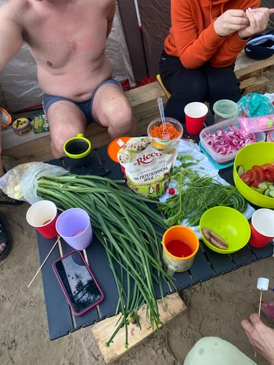 The image depicts an outdoor picnic scene on sandy ground, likely in the vicinity of Ryabinino, Russia, given the context. The focus is on a table laden with food and drinks. A man, partially visible from the chest up, is seated, wearing blue swim trunks. To his right, a person wearing an orange hoodie and black pants is engaged in a task with their hands.
On the table, a large bunch of green onions is prominently displayed, alongside a bag of Mr. Ricco mayonnaise. Various containers hold food items, including shredded carrots, chopped red onions, and a salad of tomatoes and cucumbers. A green bowl contains sliced sausages. Several disposable cups, one red and one purple, are scattered around. A yellow and black mug, and an orange mug filled with a red liquid are also present. A smartphone with a red case lies on the table, reflecting the sky and trees. In the foreground, a marshmallow is being held on a skewer. The background shows elements of a tent and wooden structures. The overall atmosphere suggests a casual outdoor gathering or meal.