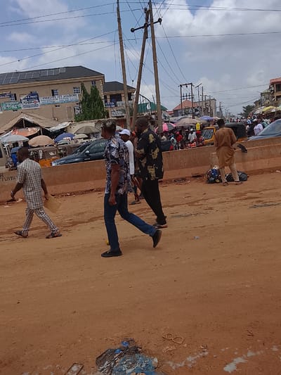 The image depicts a street scene in Katambe, Nigeria. The ground is predominantly dirt, and several people are walking along it. In the background, there is a bustling market with various stalls and people. Vehicles, including cars and auto-rickshaws (tricycles), are visible amidst the crowd. A building with solar panels on its roof and a sign reading "FIDELITY ICT ACADEMY" is also present. Overhead, electrical wires are strung between utility poles. Debris litters the foreground.