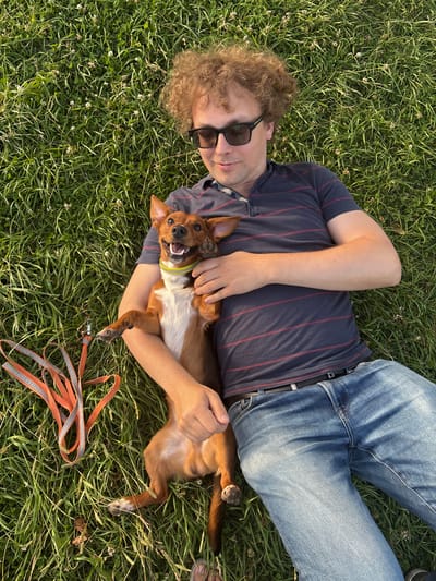 A man with curly, light brown hair and sunglasses is lying on a grassy lawn, holding a small, brown and white dog. The man is wearing a dark blue polo shirt with thin red stripes and blue jeans. The dog is lying on its back, being held by the man. Its mouth is open in a happy expression, and its tongue is visible. An orange and grey leash is lying to the left of the man and dog. The scene is illuminated by what appears to be late afternoon or early evening sunlight, casting a warm glow on the grass and subjects. The setting is a grassy area with small white flowers scattered throughout, consistent with a park or open field. There is no specific indication of the location being within the city of Gatchina, Russia, based solely on the visual information.