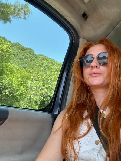 A red-haired woman wearing sunglasses and a white top is seen from the chest up, sitting in the passenger seat of a vehicle. She is looking away from the camera, towards the right. Through the open window to her left, a lush green hillside covered in trees and blue sky is visible. The interior of the vehicle, including the door panel and ceiling, are also visible. The scene appears to be a daytime outdoor setting, possibly on a road in the vicinity of Medinski Krš, Montenegro.