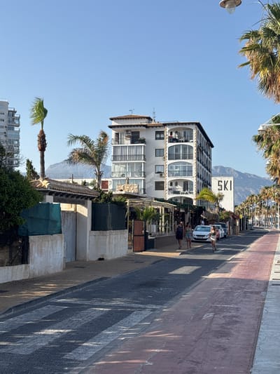 The image captures a street scene in Altea, Spain, with a prominent white building featuring arched balconies and a large "SKI" sign on an adjacent building in the background. Palm trees line the street, and mountains are visible in the distance under a clear blue sky. Several people are walking on the sidewalk, and cars are parked along the road, which has a marked pedestrian crossing. The buildings on the left appear to be older, with a textured facade and a green fence. The overall atmosphere suggests a sunny day in a coastal Mediterranean town.