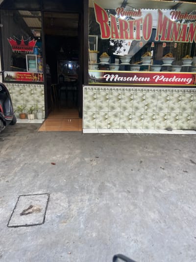 The image shows the exterior of a storefront in Kecamatan Cimahi Selatan, Indonesia. The storefront is marked with a sign that reads "Rumah Makan Barito Minan" in red lettering with the words "Masakan Padang" in black lettering on a red background below. The storefront is made of dark brown wood, with a glass window display. The window display is filled with various food items, including plates of food and bowls of food. There are two potted plants in front of the store, and a motorcycle parked in the foreground. The ground in front of the store is made of concrete. The motorcycle is facing the camera and is partially obscured by the right edge of the image. The ground is light grey, with a drainage grate. The front of the building is tiled in an alternating pattern of white, grey, and tan. The door to the store is open, and the interior of the store is visible. The interior has a grey and white tiled floor, two stools, and a couple of tables. The location of the store can be determined by the coordinates provided: -6.932, 107.564. The scene appears to be abandoned or unoccupied. No people are present, and there are no other signs of activity.