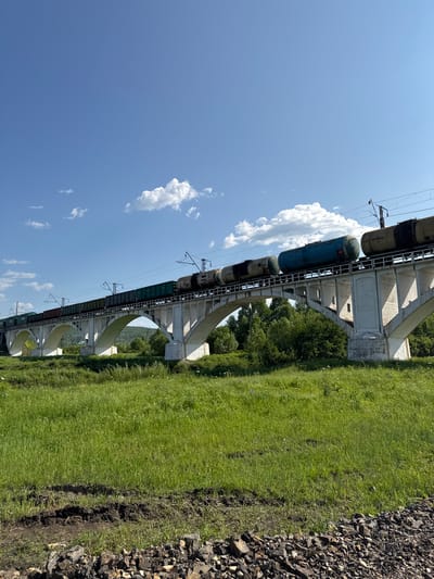 A train is crossing a viaduct in Skovorodino, Russia. The train consists of multiple cars, including tankers and cargo containers, all of which are visible on the elevated track. The viaduct itself is a substantial concrete structure with multiple arched supports, suggesting it spans a significant distance or height. The surrounding landscape is green and lush, with a grassy field on the right side of the frame and a line of trees in the background, partially obscured by the viaduct. The sky is a clear blue with a few scattered white clouds, indicating a sunny day. The time of day appears to be afternoon, judging by the lighting and shadows. There is a pile of gravel or rocks in the foreground on the right, adjacent to the grassy field. Some text is visible on the train cars, but it is not clear enough to be read. The overall scene depicts a functional railway infrastructure integrated into a natural, rural environment.