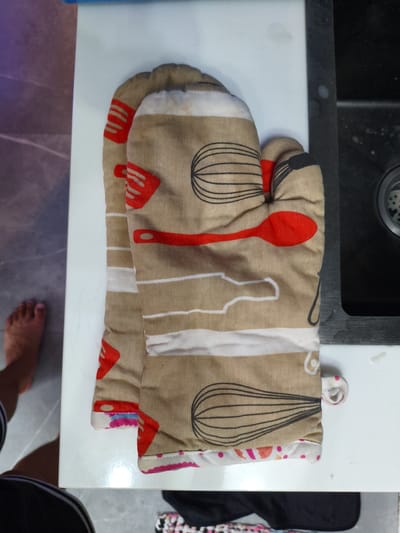 This is a top-down, eye-level, medium shot of a pair of oven mitts with a kitchen utensil pattern lying on a white countertop. The mitts are tan with brown and white stripes, and feature illustrations of whisks, spatulas, and spoons in red and black. A foot is visible on the lower left of the frame, suggesting a person is present but not fully in view. The background includes a dark sink on the right and a sliver of a tiled floor on the left. The overall environment appears to be a domestic kitchen setting. There is no indication of the specific location within Cavite City, Philippines.